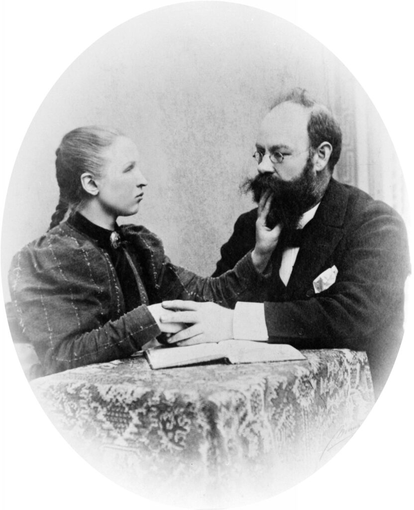 Ragnhild Kaata (to the left) sitting behind a table with Elias Hofgård. Ragnhild's left hand is placed on Hofgård's chin with two fingers touching his lower lip. Her vacant hand rests in Hofgård's left hand on the table. The two are facing each other. On the table lies an open book.