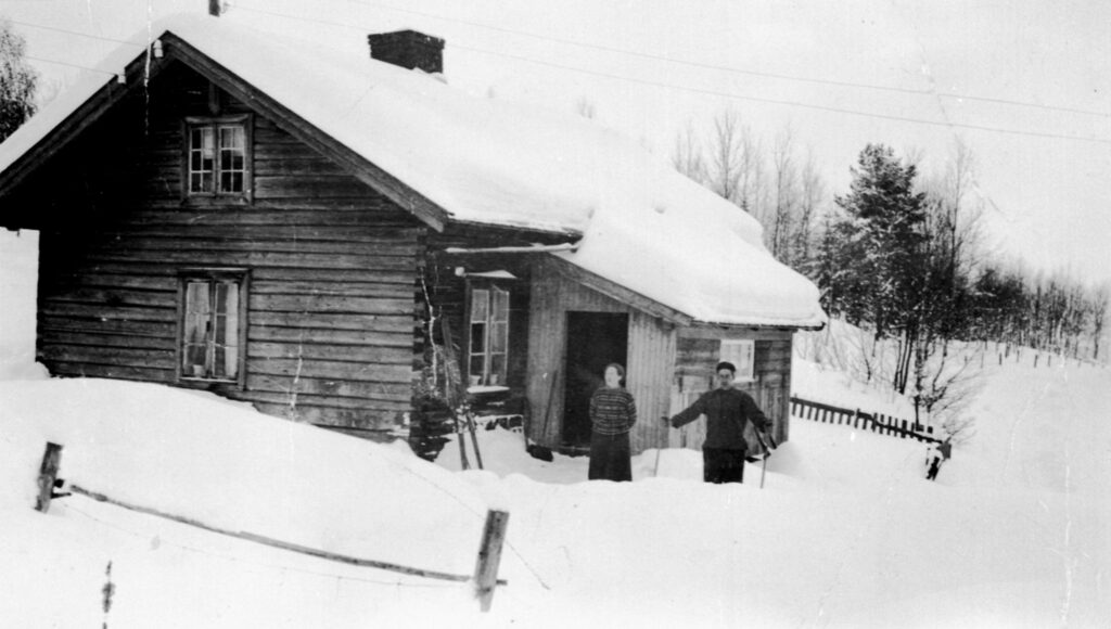 Example of a tenant farm cottage, early 20th century. Small and simple two-story timber house with no paint. The thick layer of snow on the ground reaches up to the windows at ground floor. A 20 inch layer of snow covers the roof. A man and a woman standing outside the entrance, the man in skiing outfit.