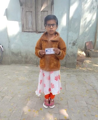 A female child with short hair and glasses is standing in a street holding an ID card.