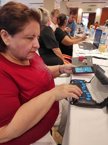A woman in a red top is using a braille line with her right hand, whilst with her left hand she holds her phone.