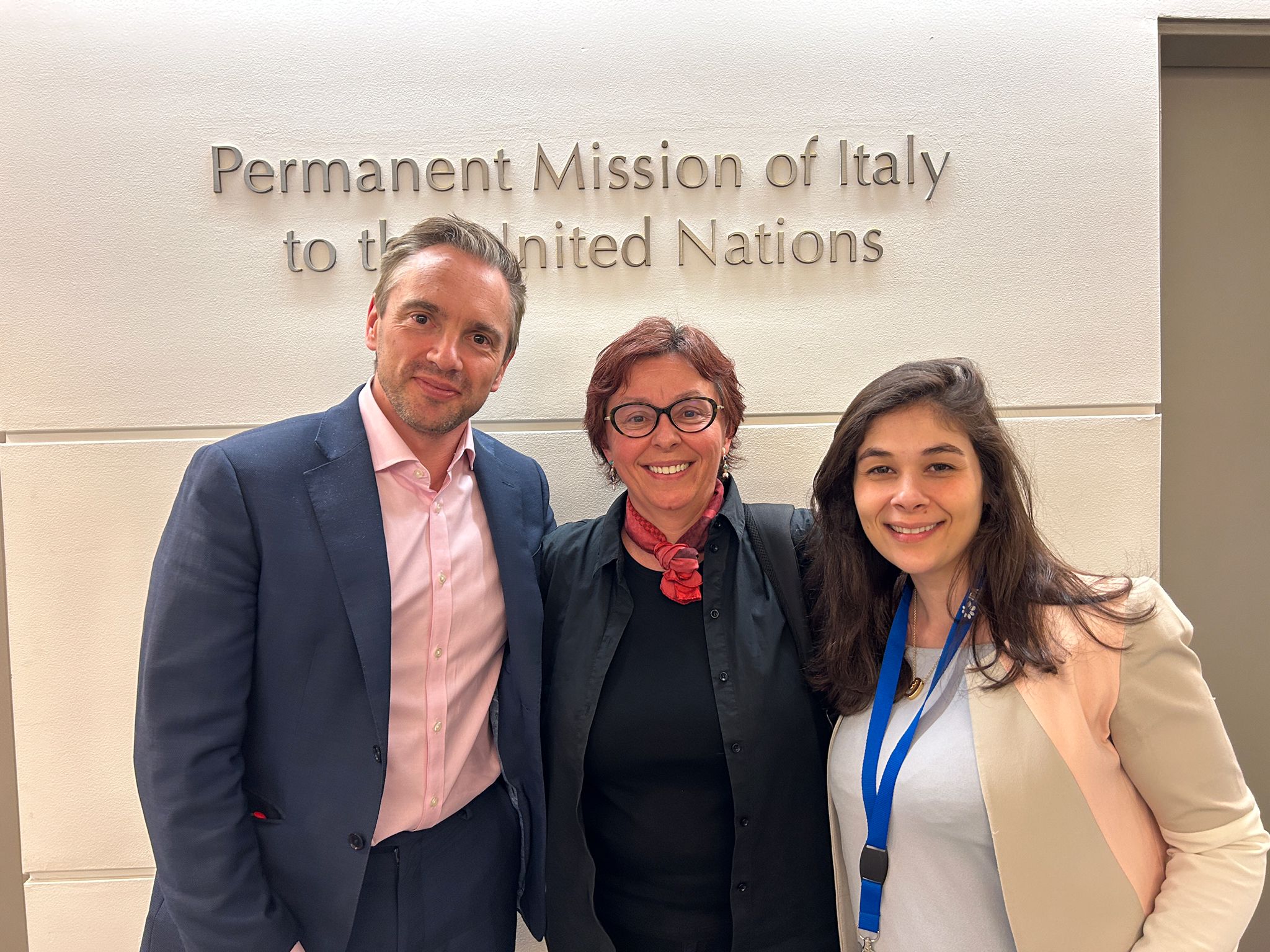Two women and a men pose in front of a sign that reads "Permanent Mission of Italy to the United Nations"