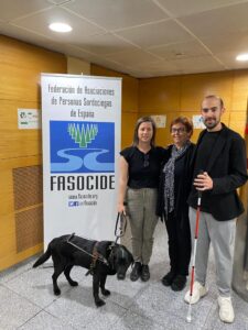 Two women and a man stand next to a banner that reads "FASOCIDE". One is assisted by a black guide dog and another is holding the red and white cane