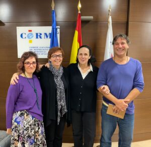 Three women and a man pose for the camera standing up. Behind them, three flags and a sign that reads "CERMI"