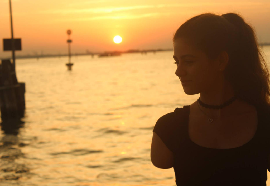 A woman is looking sideways and shows an amputated arm. In the background, the sea, and a sunset
