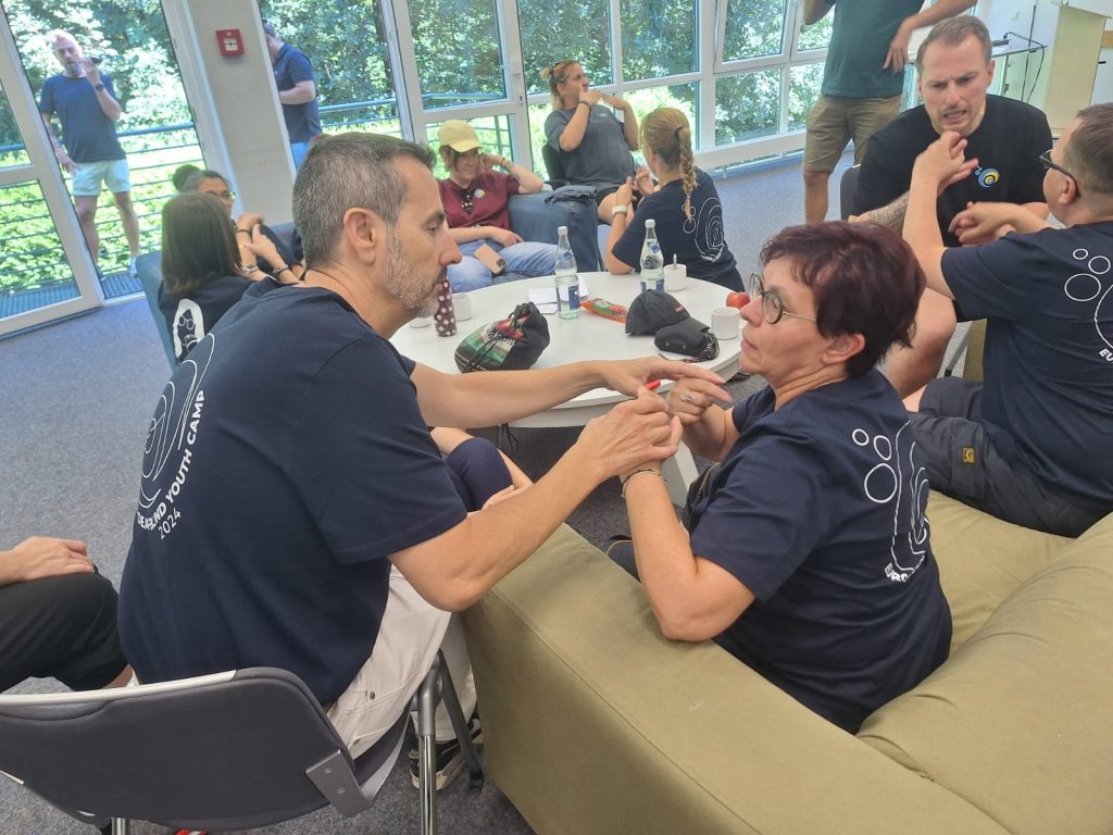 Frank and Sanja are sitting and communicating with each other using tactile sign language. In the background, other people are visible, all wearing the same dark blue t-shirt.