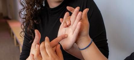 Close-up of two people engaging in tactile communication, with one person gently touching and gesturing on the other's hand. The individuals' faces are partially out of frame