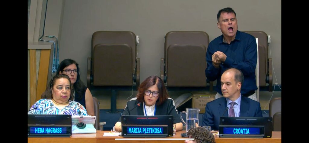 Two women and a man sat at a table, behind them, a woman wearing glasses and a man standing who appears to be signing