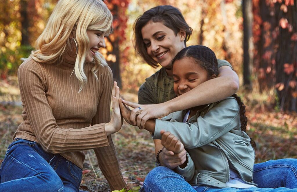 Three people—two adults and one child—sit outdoors in a wooded area during autumn, smiling and interacting warmly. One adult gently guides the child's hands in a tactile gesture