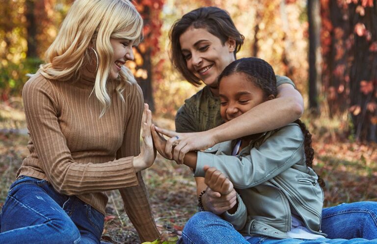 Three people—two adults and one child—sit outdoors in a wooded area during autumn, smiling and interacting warmly. One adult gently guides the child's hands in a tactile gesture