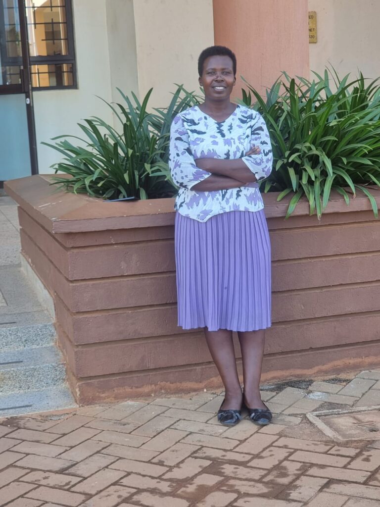 Juliet is standing straight with her hands crossed at the wrists, smiling. She is wearing a purple dress with a white top patterned with purple and black markings, and a plain purple skirt with even tacking that reaches just below her knees. She is also wearing black closed-toe shoes.